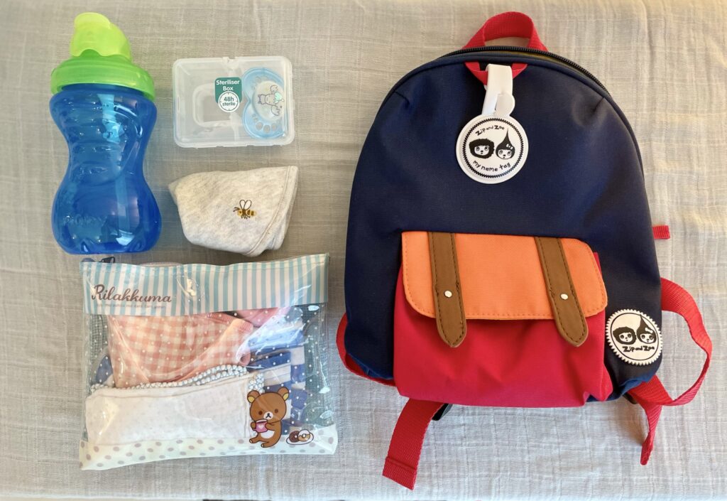 Showing the contents of Zip and Zoe mini backpack in navy colourblock - Nuby water bottle, pacifier, bib, clothes